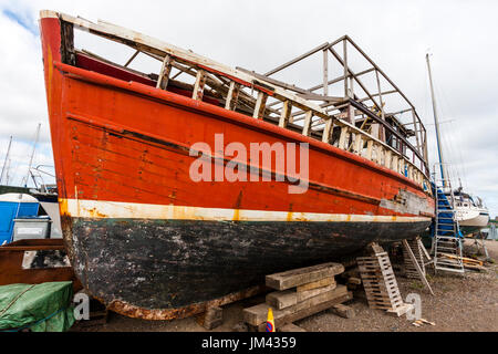 Ehemalige Fischerboot, rot Rumpf, unterstützt durch Hölzer auf dem Land und in der Tat - Reparatur und Verwitterung. Teile von Hull durchlöchert, Farbe in Orten entfernt. Stockfoto