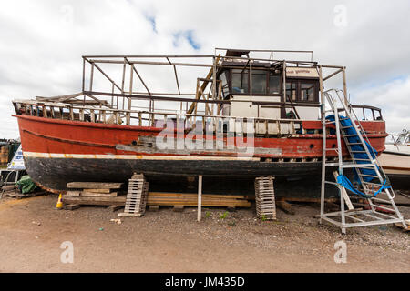 Ehemalige Fischerboot, rot Rumpf, unterstützt durch Hölzer auf dem Land und in der Tat - Reparatur und Verwitterung. Teile von Hull durchlöchert, Farbe in Orten entfernt. Stockfoto