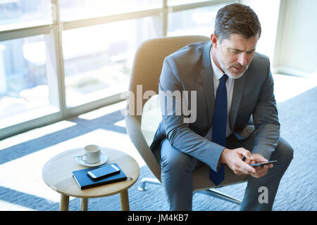 Vogelperspektive Blick auf eine ausgereifte Business executive sitzen in einem Büroraum mit großen Fenstern, die eine SMS-Nachricht auf seinem Handy lesen Stockfoto