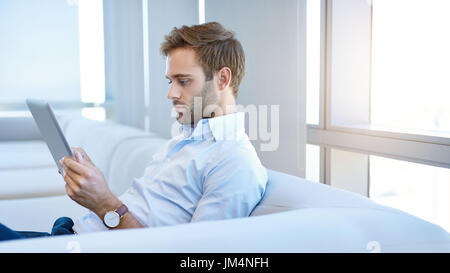 Stilvolle junger Mann sitzt auf einer modernen Couch ernsthaft lesen Informationen über ein digital-Tablette, mit Licht, das durch große Fenster hinter ihm Stockfoto