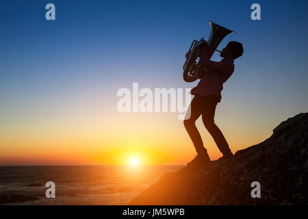 Silhouette der Musiker spielt die Trompete auf felsigen Küste während des Sonnenuntergangs. Tuba-Instrument. Stockfoto