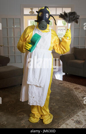 Reife Frau in gelb Haz-Mat Suir und Gasmaske stehend im Wohnzimmer mit Schwamm und Staubwedel. Stockfoto