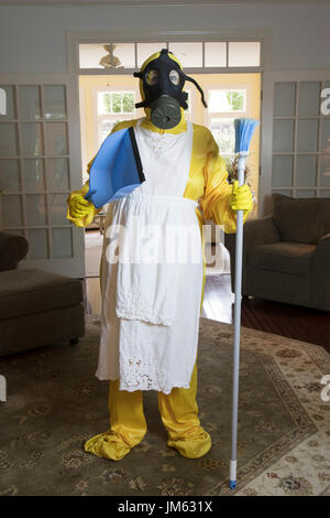 Reife Frau in gelb Haz-Mat Suir und Gasmaske im Wohnzimmer mit Besen und Staub Pfanne stehen. Stockfoto