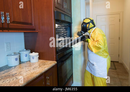 Reife Frau in gelb Haz-Mat Suir und Gasmaske Türöffnung zu Ofen in der Küche zu Hause, Stockfoto