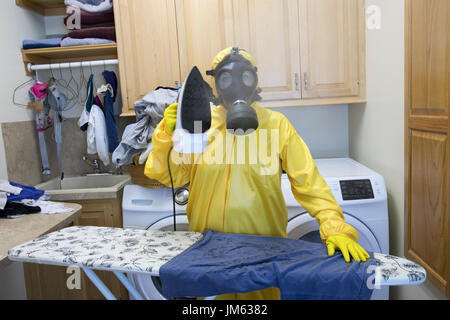 Reife Frau in gelb Haz-Mat Suir und Gasmaske Bügeln Hemd an Bord mit Dampfbügeleisen. Stockfoto