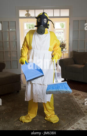 Reife Frau in gelb Haz-Mat Suir und Gasmaske im Wohnzimmer mit Besen und Staub Pfanne stehen. Stockfoto