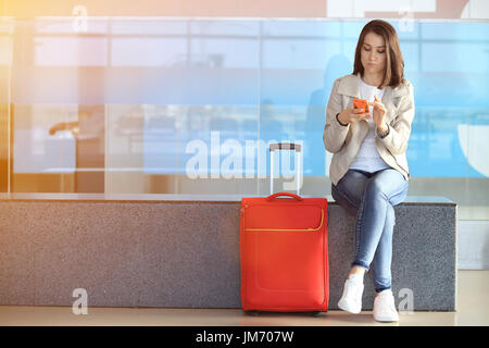 Mädchen mit Smartphone sitzen in der Nähe von roten Koffer im Flughafen. Hübsches Mädchen warten auf den nächsten Flug. Stockfoto