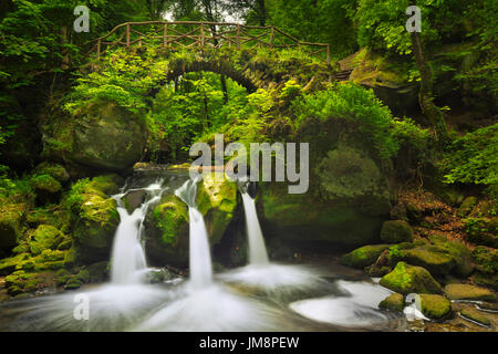Eine steinerne Brücke über einen Fluss mit einem kleinen Wasserfall in der Nähe von Müllerthal, Luxemburg. Stockfoto