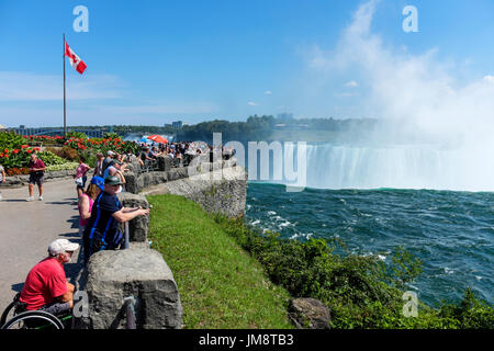 Besucher zu den Niagara Fällen sehen die kanadischen Horseshoe Falls aus Sicht. Gischt der Wasserfälle liegt in der Luft an einem sonnigen Tag. Kanadische Flagge. Stockfoto