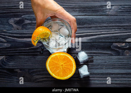 Bartenders Hände am Werk-Nahaufnahme. Vorbereitung für ein erfrischendes Getränk mit Eiswürfel und Zitrusfrüchten. Draufsicht auf einen dunklen hölzernen Balken Stockfoto