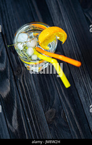 Gin Tonic mit Eis, verziert mit einer Orangenscheibe und Rosmarin. Cocktail mit Orangenscheibe und zwei Strohhalme auf dunklen Holztisch Stockfoto