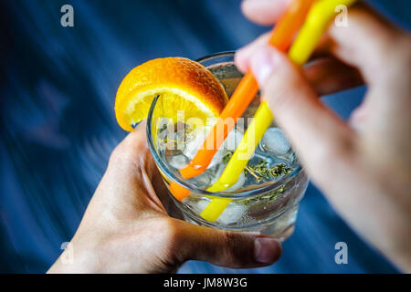 Gin Tonic mit Eis, verziert mit einer Orangenscheibe und Rosmarin. First-Person-Ansicht ein alkoholfreies Getränk in eine Glasschale mit ein paar Strohhalme Stockfoto