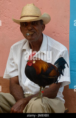 Ein Mann sitzt mit seinem Hahn auf seine Knie und raucht eine Zigarre. In Trinidad, Kuba gedreht. Stockfoto