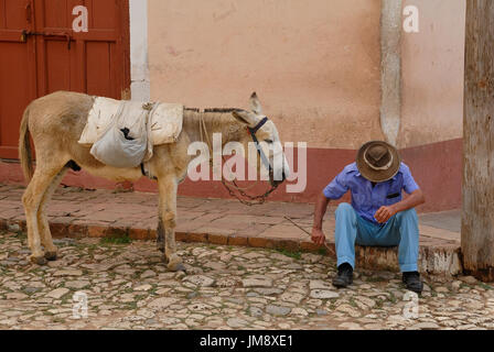 Ein Mann sitzt und ruht auf dem Bürgersteig in einer der Straßen in den alten, kolonialen, Teil von Trinidad in Kuba. Seinen Esel wartet geduldig. Stockfoto