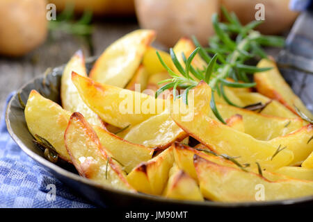 Gebackene Kartoffelspalten mit Rosmarin in einer Eisenpfanne, rohen Kartoffeln im Hintergrund Stockfoto