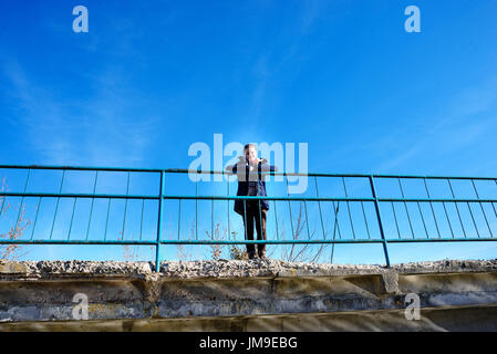 Auf einer Betonbrücke ist eine ruhige, glückliche Frau liebt Wandern. Eine Frau im Hintergrund des blauen Herbsthimmel sieht irgendwo in der Ferne. Stockfoto