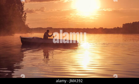 Ein junger Mann fährt ein Boot auf einem See während einer goldenen Sonnenuntergang. Bild der Silhouette, Sonnenuntergang. Mann ein Ruderboot im Gegenlicht der Sonne. Contre-Jour, backli Stockfoto