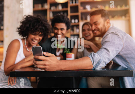 Junge Männer und Frauen am Café-Tisch sitzen und nehmen ein Selbstporträt mit Handy. Gemischtrassigen Gruppe von Freunden nehmen Selfie auf Smartphone bei c Stockfoto