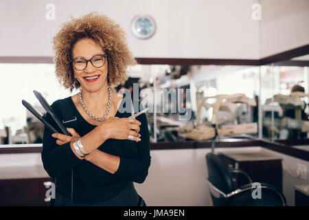 Professionelle Friseur hält einen Haarglätter und Schere. Frau Friseur in fröhliche Stimmung im Salon. Stockfoto