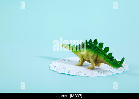 Ein Dinosaurier Stegosaurus auf eine weiße Spitze Papier Deckchen mit einem lebhaften Türkis Hintergrund. Minimale ausgefallenen Still-Life Fotografie Stockfoto