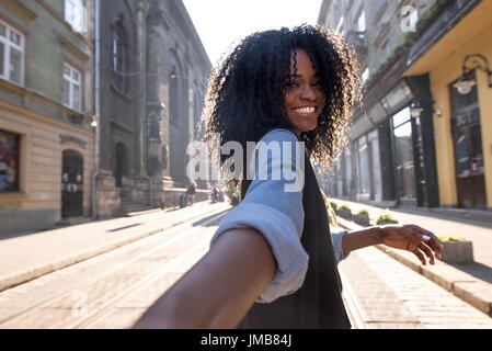 Outdoor-Porträt von attraktiven dunkelhäutige Frau mit lockigen Haaren herumlaufen Stadt am sonnigen Tag, Blick in die Kamera mit freudigen hübsches Lächeln schönes Wetter genießen Stockfoto