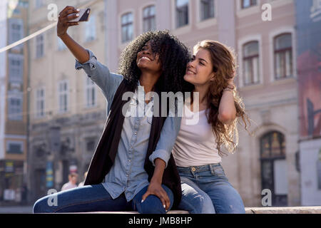 Zwei Freunde machen Selfie, lächelte. Frauen tragen legere Kleidung. Eine Frau ist schwarz. Schöne Architektur Hintergrund. Stockfoto