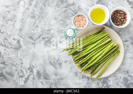 Bündel von rohen Spargel auf Teller, Salz, Pfeffer und Olivenöl auf hellem Hintergrund. Ansicht von oben Stockfoto