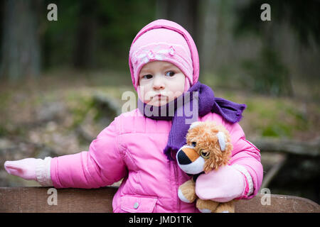 Mädchen mit braunen Augen und rosa Jacke steht auf der Bank im Park und haben ein Spielzeug Hund Welpen Stockfoto