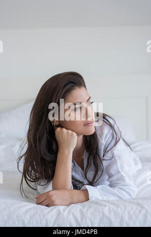 Junge Frau am Bett liegen und trägt ein weißes Hemd. Sie stützte sich auf Ellbogen und wegsehen. Stockfoto