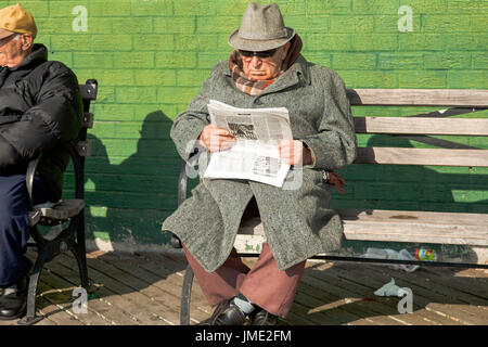 Ein Mann im Mantel und Hut liest eine Russische Zeitung in Brighton Beach, New York City. Er sitzt auf einer Bank vor einer grünen Wand. Stockfoto