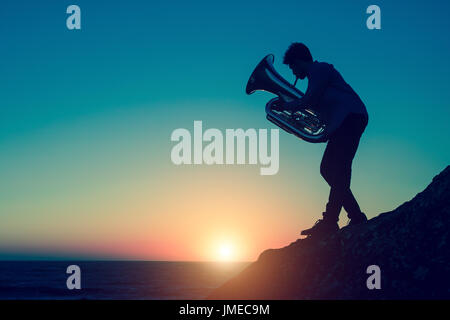 Silhouette der Musiker spielt die Tuba an felsigen Küste bei traumhaften Sonnenuntergang. Trompete. Stockfoto