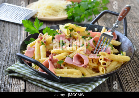 Nudeln mit gekochtem Schinken und Speck-Bits, Käse und Eiern in einer Pfanne, geriebenem Käse im Hintergrund Stockfoto