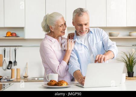 Bild des Lächelns Reifen liebenden paar Familie stehen in der Küche in der Nähe von Tee und Gebäck mit Laptopcomputer. Auf der Suche zur Seite. Stockfoto