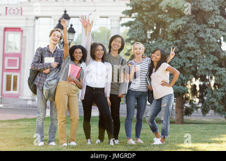 Bild der multiethnische Gruppe von jungen fröhlichen Studenten stehen und winken im Freien. Blick in die Kamera. Stockfoto
