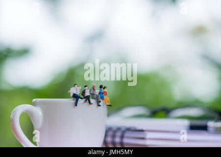 Miniatur Geschäftsmann Buch lesen und sitzen auf der Tasse Kaffee mit Brille und Notebook Hintergrund über als Business Konzept. Stockfoto