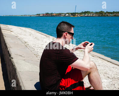 Mann mit Sonnenbrille sitzt in der Nähe der Uferpromenade mit Smartphone Stockfoto
