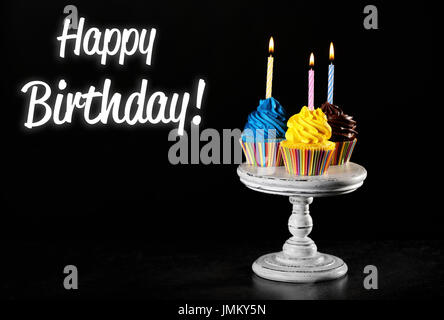 Leckere Muffins mit Kerzen und Text HAPPY BIRTHDAY auf schwarzem Hintergrund Stockfoto