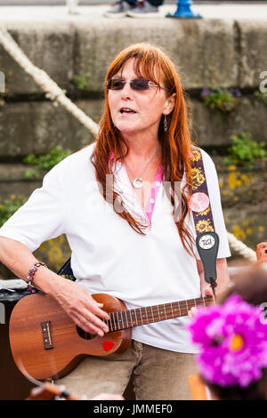 Reifen kaukasischen Frauen, 50 s, stehen im Freien spielen Ukulele. Konfrontiert, aber Weg suchen, trägt eine Sonnenbrille. Stockfoto