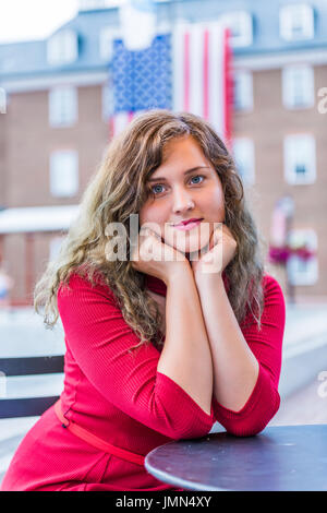 Junge Frau im roten Kleid sitzt lächelnd mit amerikanischer Flagge der Vereinigten Staaten im Hintergrund Stockfoto