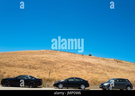 Eine Linie von drei Autos parkt in der Nähe von einem Stroh bedeckten Hügel, mit einem strahlend blauen Himmel oben, im See Chabot Regional Park, ein Regionalpark East Bay in Castro Valley, Kalifornien, 4. Juli 2017. Stockfoto