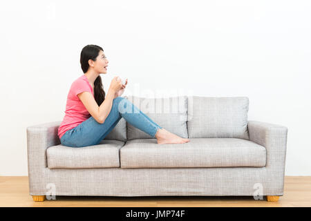 fröhlich lächelnde Frau sitzend auf Sofa entspannen, mit Blick auf weißem Hintergrund und hält heiße Tasse Becher Tagträumen in Holzboden. Stockfoto