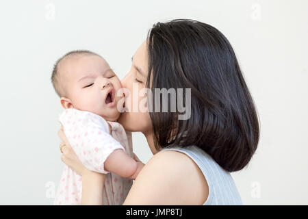 Porträt der glückliche asiatische Mutter küssen und umarmen ihre niedlichen kleinen Baby auf grauem Hintergrund Stockfoto