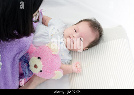 Porträt von einem kleinen entzückenden Säugling Baby Mädchen mit Bär Puppe auf Rücken auf dem Bett liegend und auf der Suche nach ihrer Mutter Stockfoto