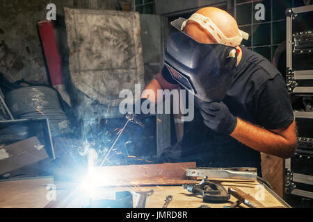 Ein glatzköpfiger Mann in einem schwarzen T-Shirt schafft mit seinen eigenen Händen ein mettalical Produkt- und schweißt die Metall mit einem schweissgerät in einer dunklen Umgebung, in der Stockfoto