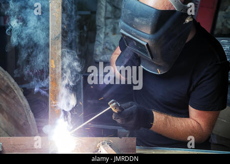 Ein kahler, starken Mann in Schwarz Arbeitskleidung Schweißnähte ein Metall Produkt mit Arc Welding Machine im Lager, einen dunklen Hintergrund, viel Rauch, hell Stockfoto