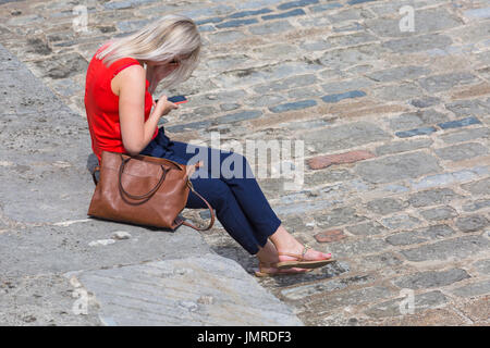 Junge Frau sitzt auf der Hafenmauer mit Handy bei Lyme Regis, Dorset im Juli