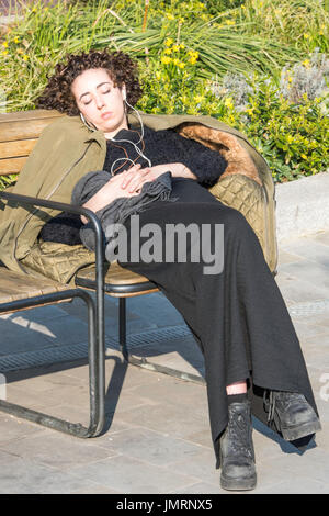 Eine junge Frau, hören von Musik in der Sonne liegend auf einer Bank in ihrer Arbeitskleidung. Stockfoto