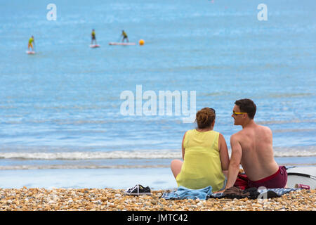 Pärchen sitzen am Strand von Lyme Regis mit Paddleboardern in der Ferne, Lyme Regis, Dorset UK im Juli Stockfoto