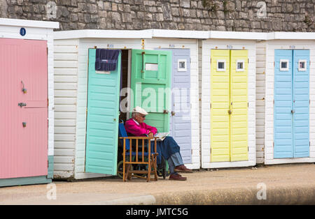 Älterer Mann sitzt lesen Zeitung am Strandhütte auf Promenade bei Lyme Regis, Dorset im Juli Stockfoto