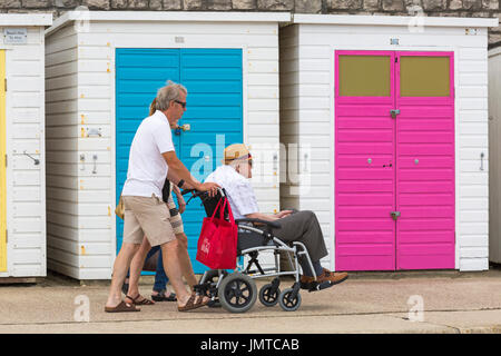 Paar treibt älteren Mann im Rollstuhl Promenade vorbei an bunten Strandhäuschen bei Lyme Regis, Dorset im Juli Stockfoto